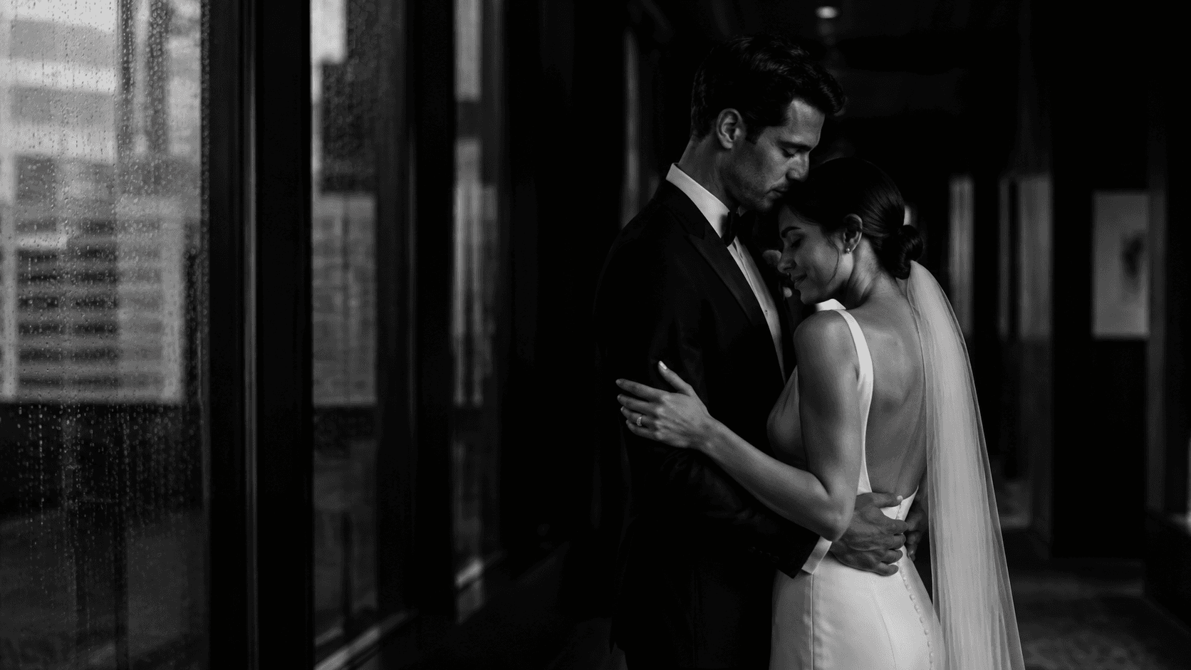 Newlyweds sharing a quiet embrace in a rain-darkened hotel corridor